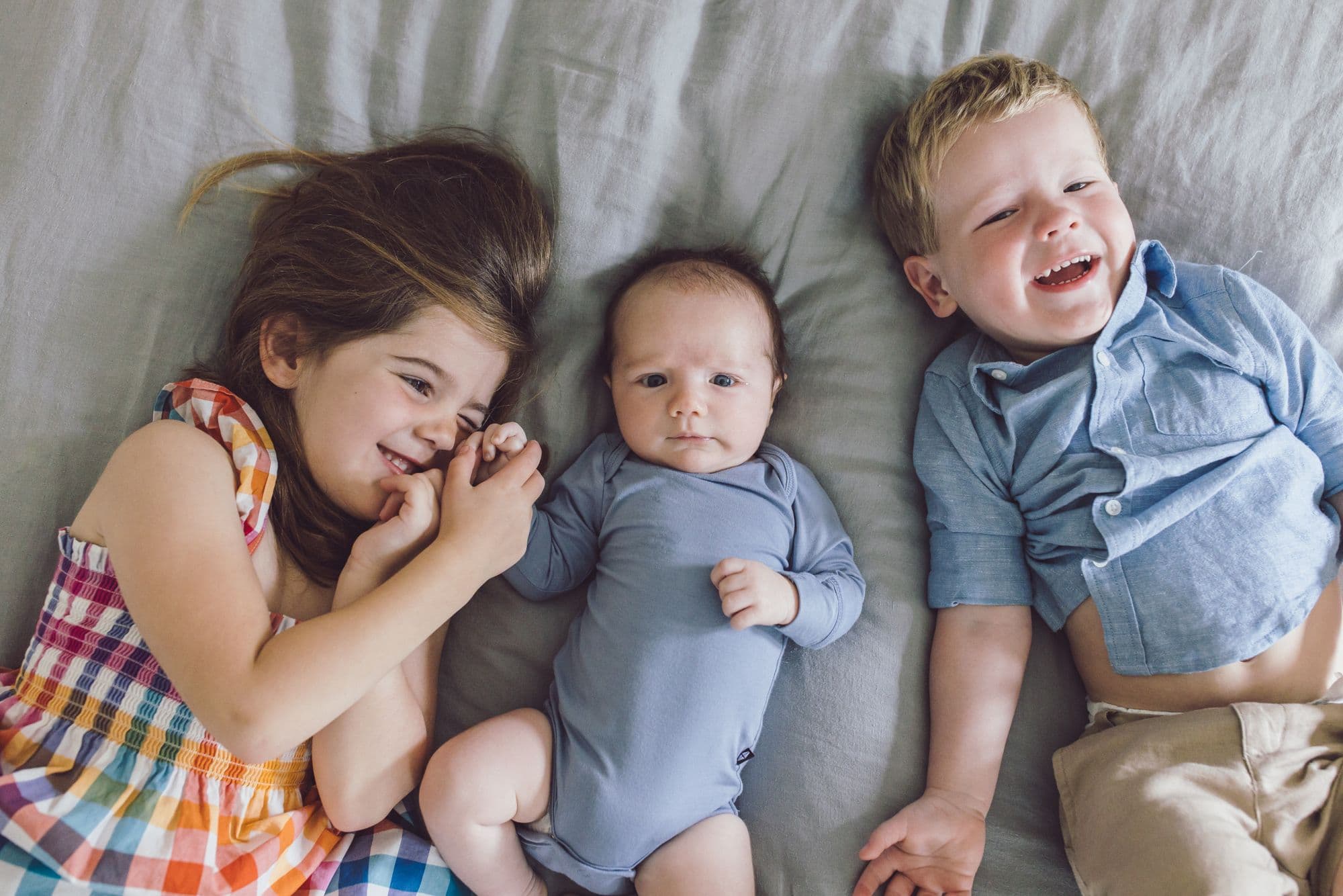 Three siblings lying side by side on a bed, the newborn between an older sister and brother