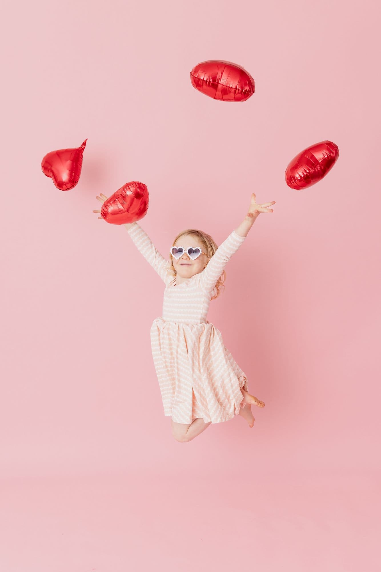 Girl in heart-shaped sunglasses jumping to catch red heart balloons against a pink studio backdrop