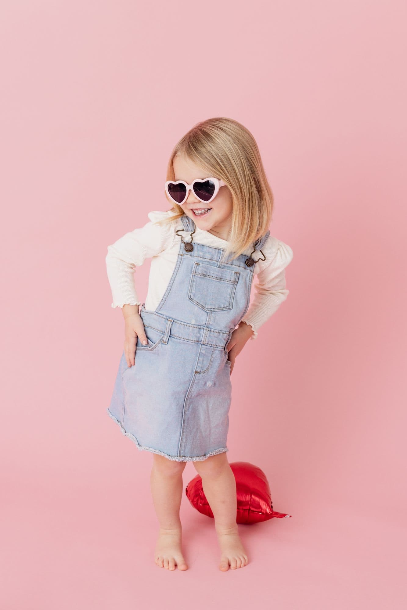 Girl in a denim pinafore and heart-shaped sunglasses against a pink backdrop