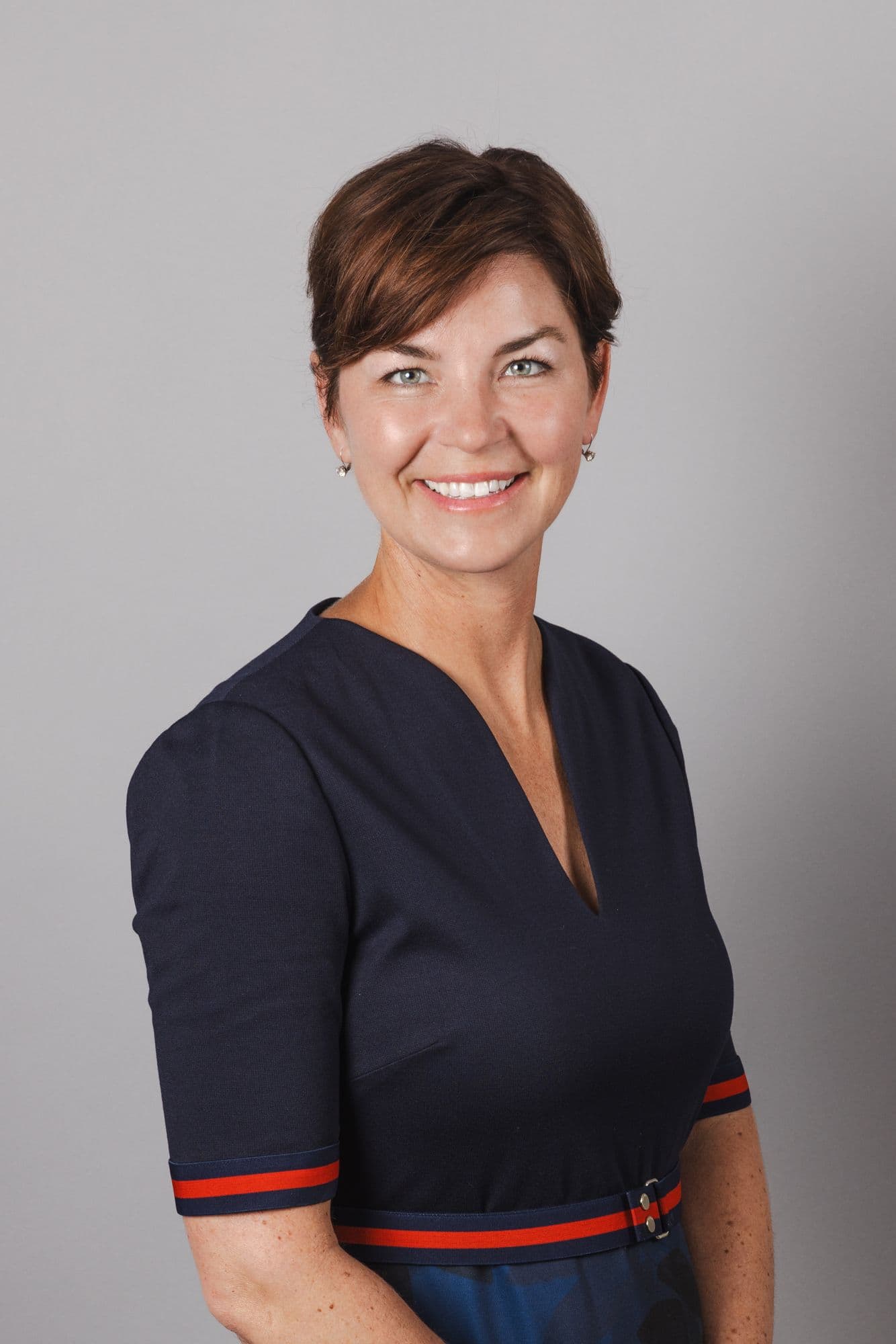 Woman in a navy dress with red trim, smiling in a studio business headshot against a gray background
