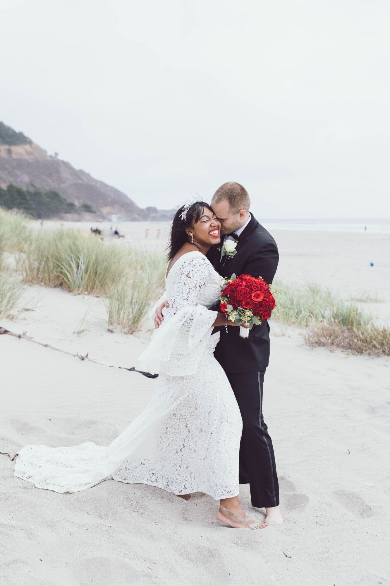 Newlyweds laughing together on the sand, the bride holding red roses