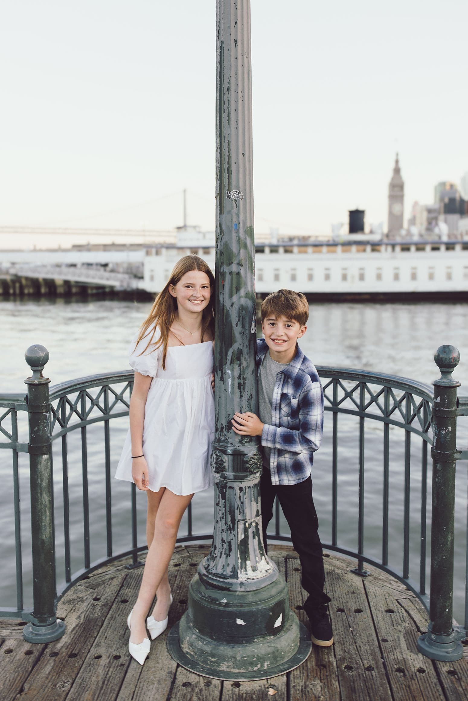 Sister and brother standing at a lamppost on a pier with the city skyline behind