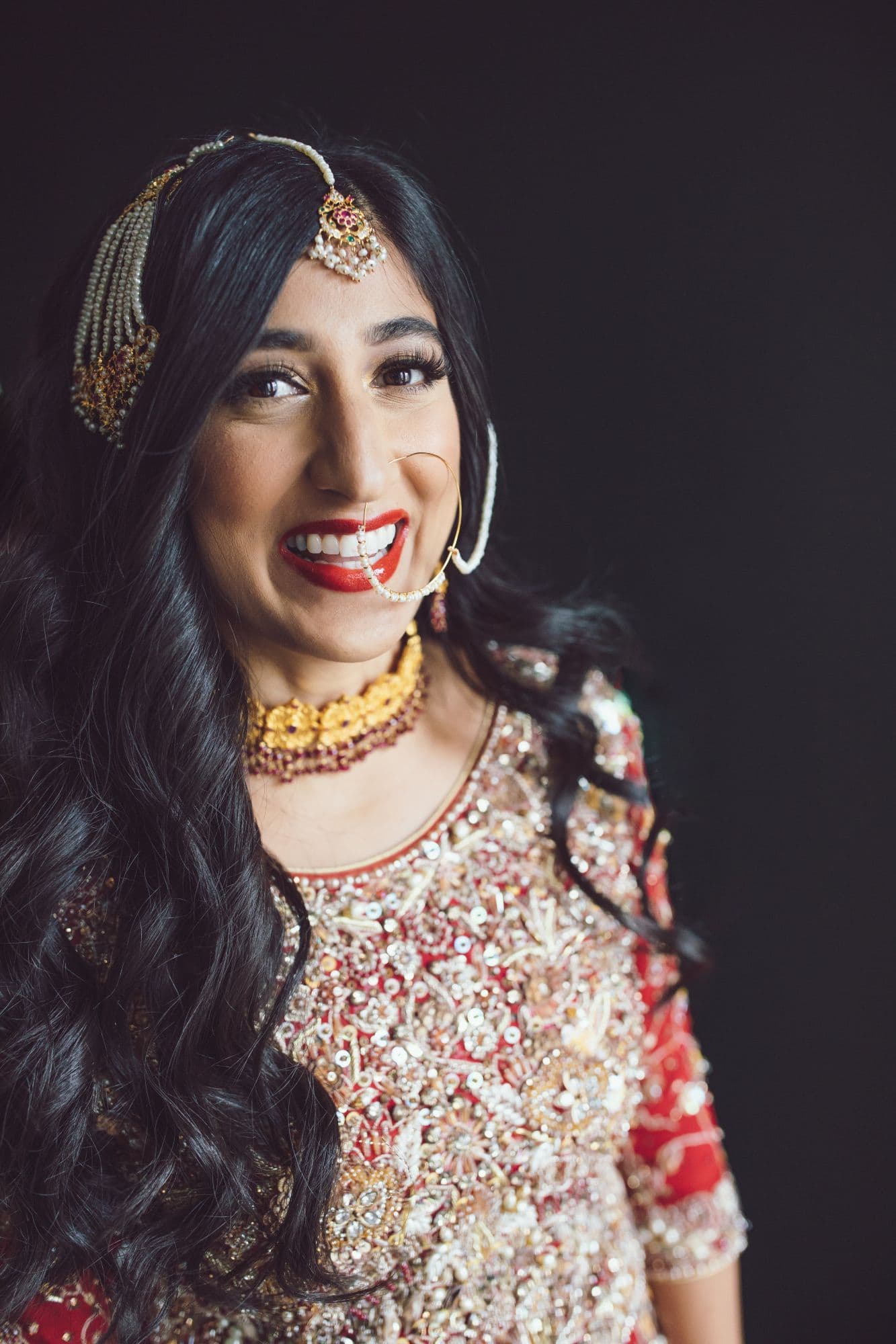 Bride in gold-embroidered bridal wear and a maang tikka, laughing, against a dark studio backdrop