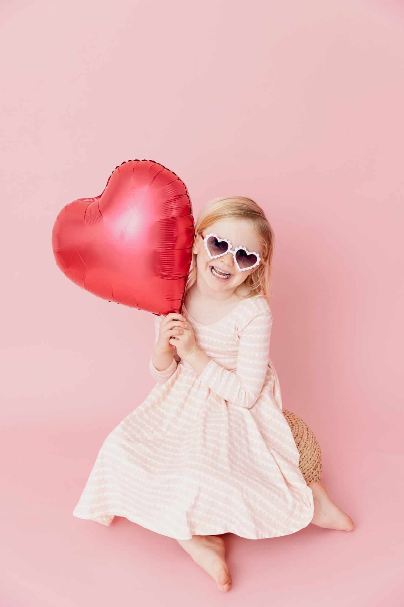 Girl in a pink dress holding a large red heart balloon against a pink ground