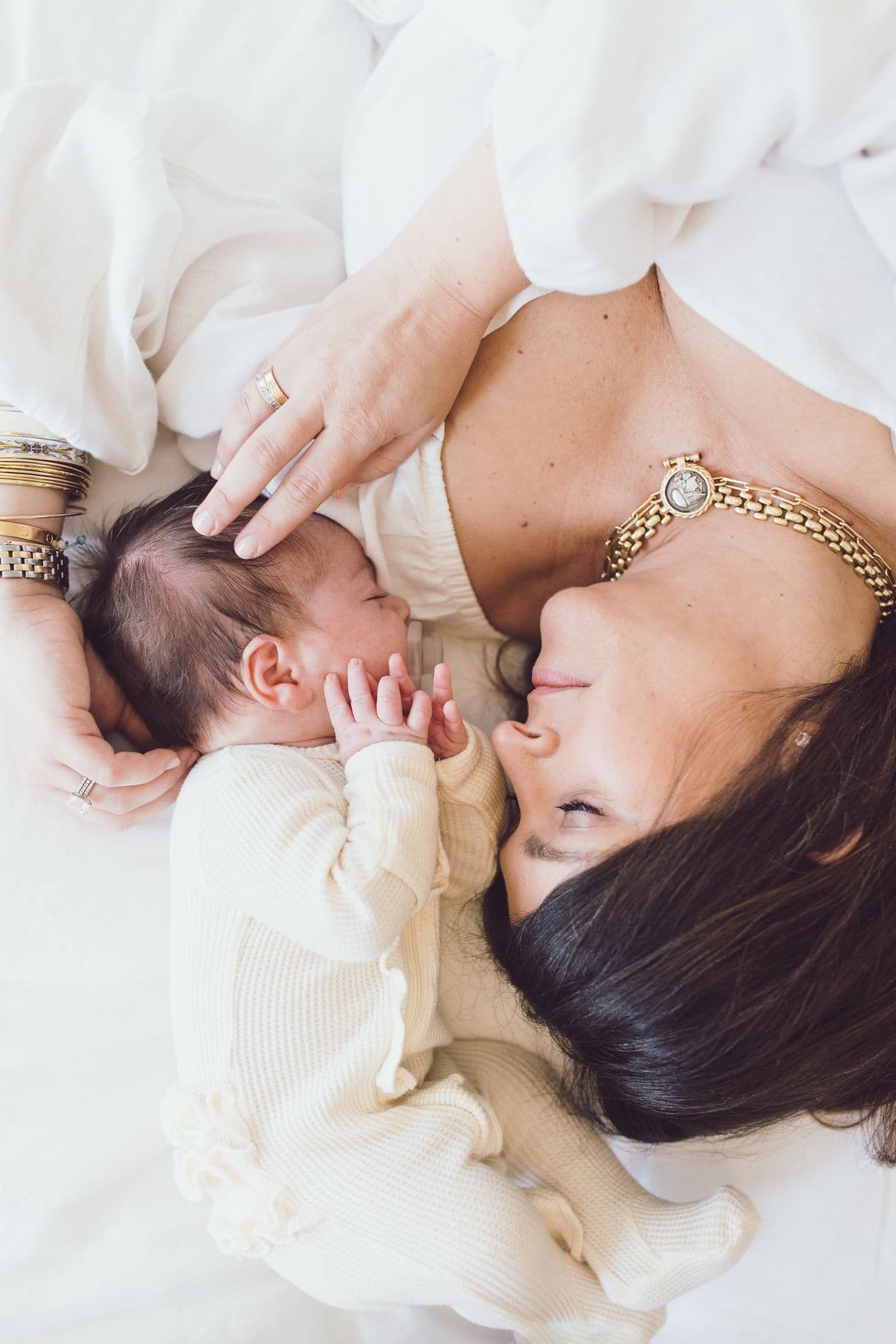 Mother lying on white bedding face to face with her newborn, one hand resting on the baby's head