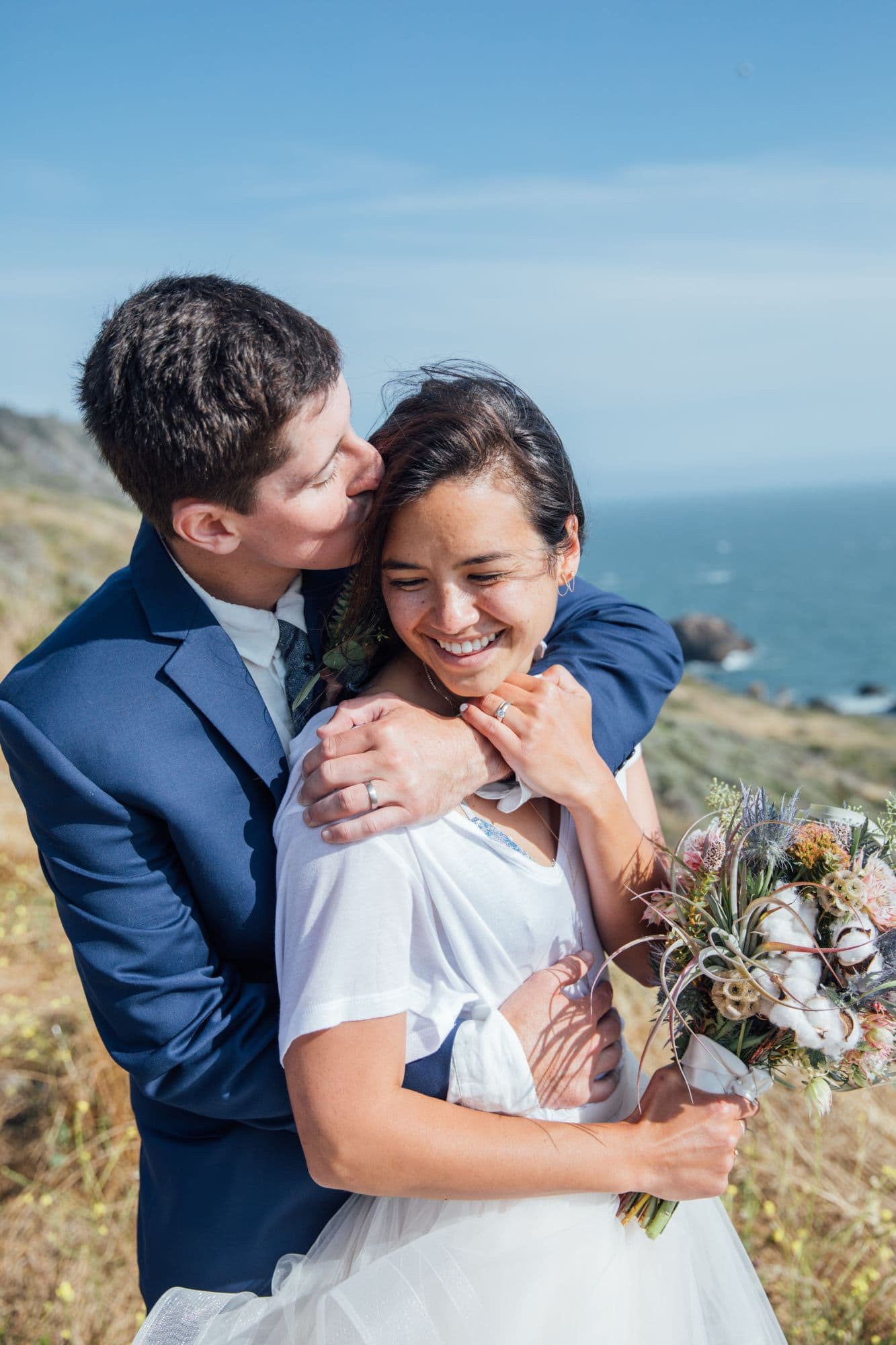 Newlyweds embracing on a coastal bluff, one holding a wildflower bouquet