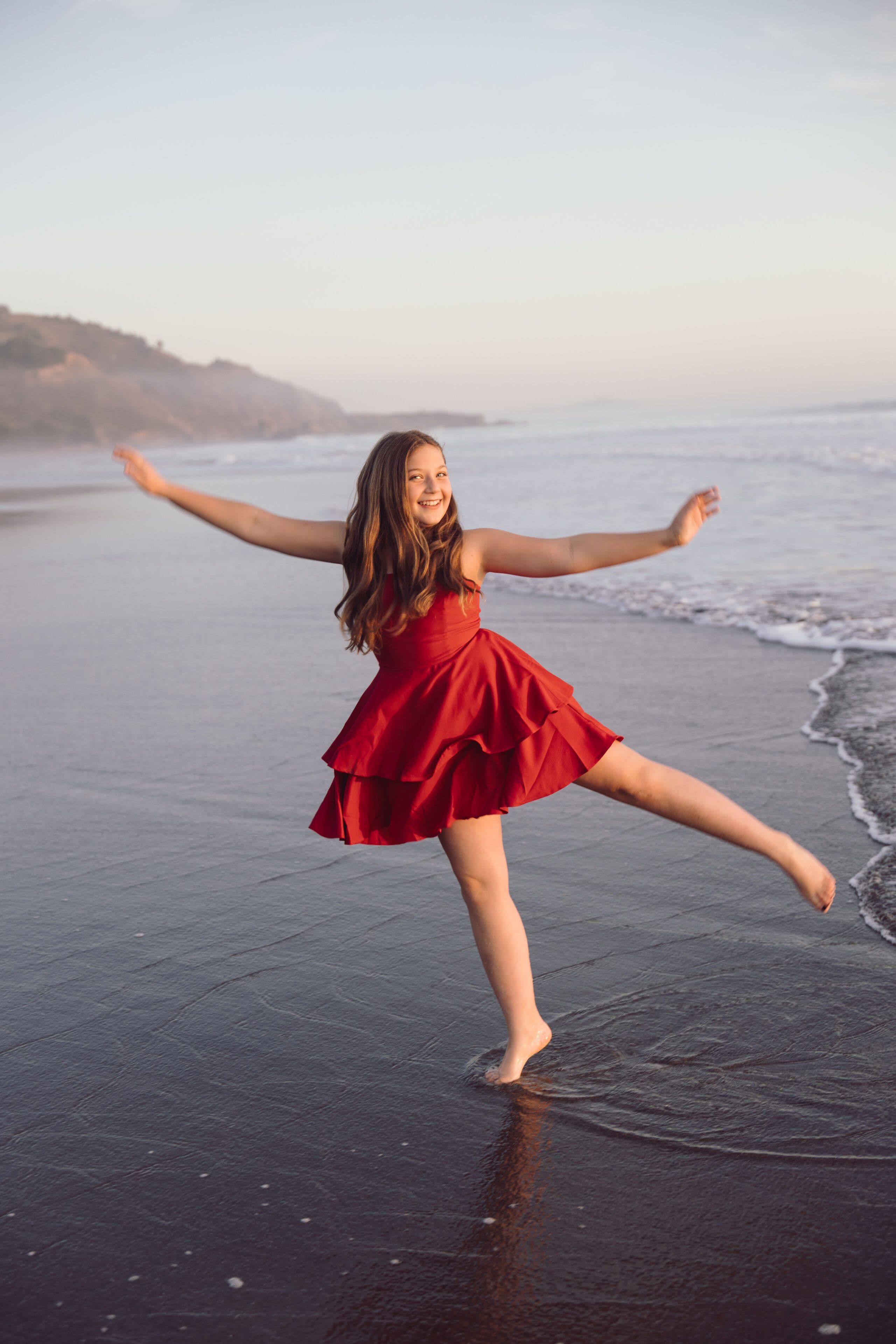 Girl in a red dress spinning barefoot at the edge of the surf at sunset