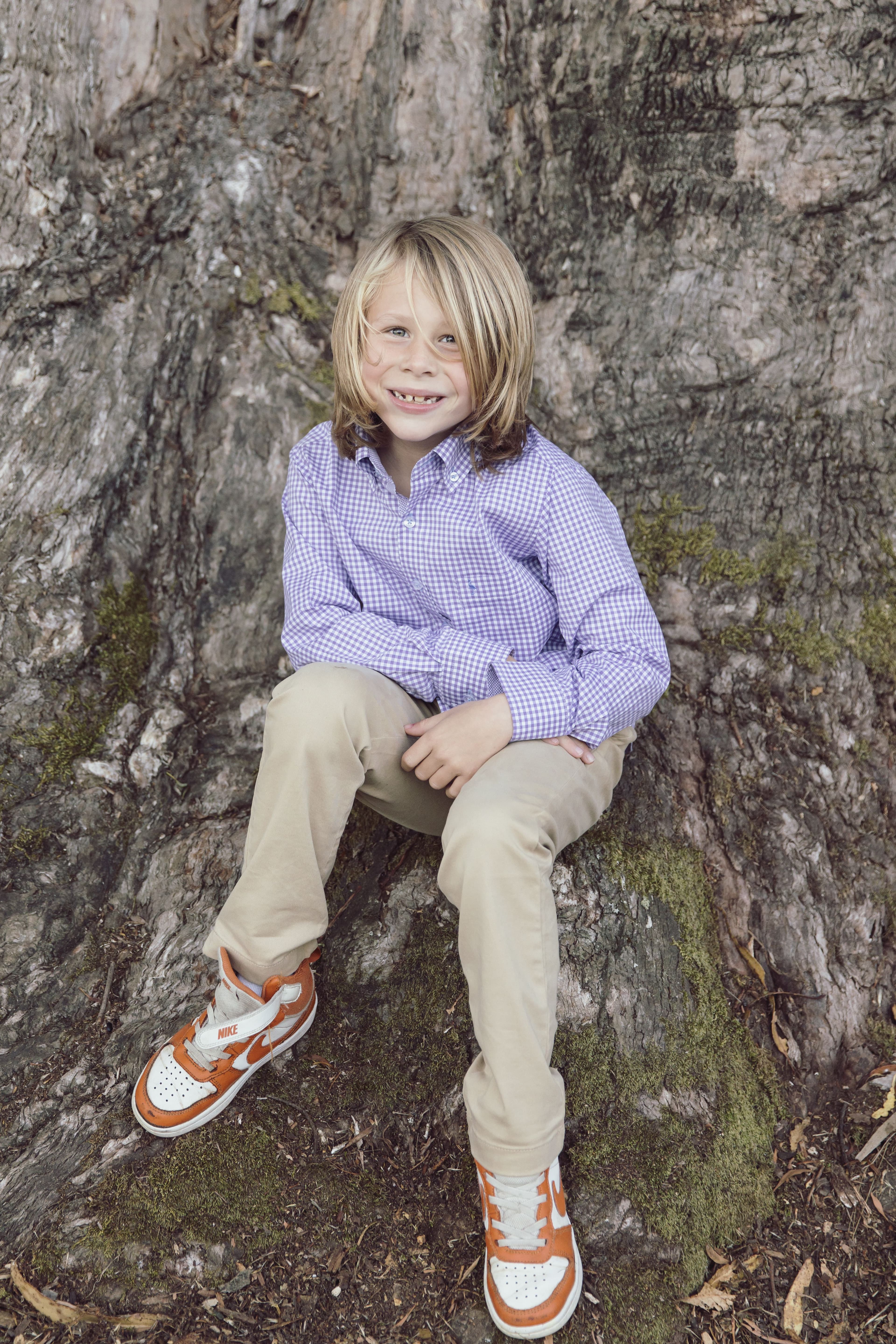 Boy in a purple gingham shirt sitting against the trunk of a large tree