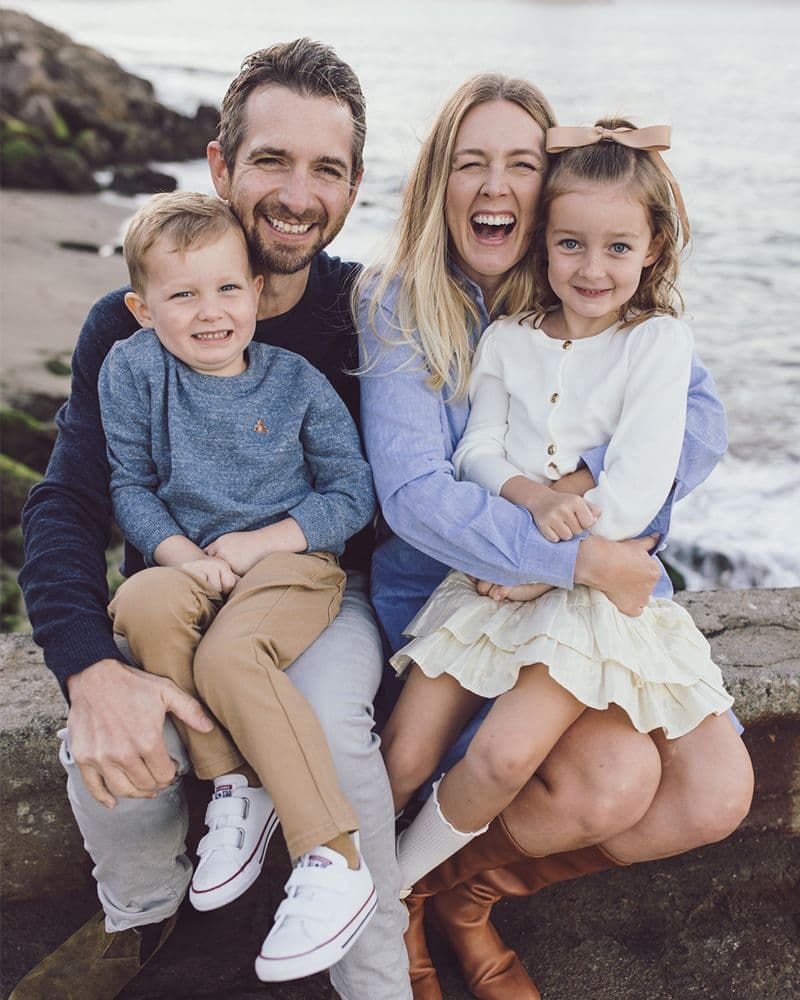 Family of four sitting close together on a low seawall by the water, laughing at the camera