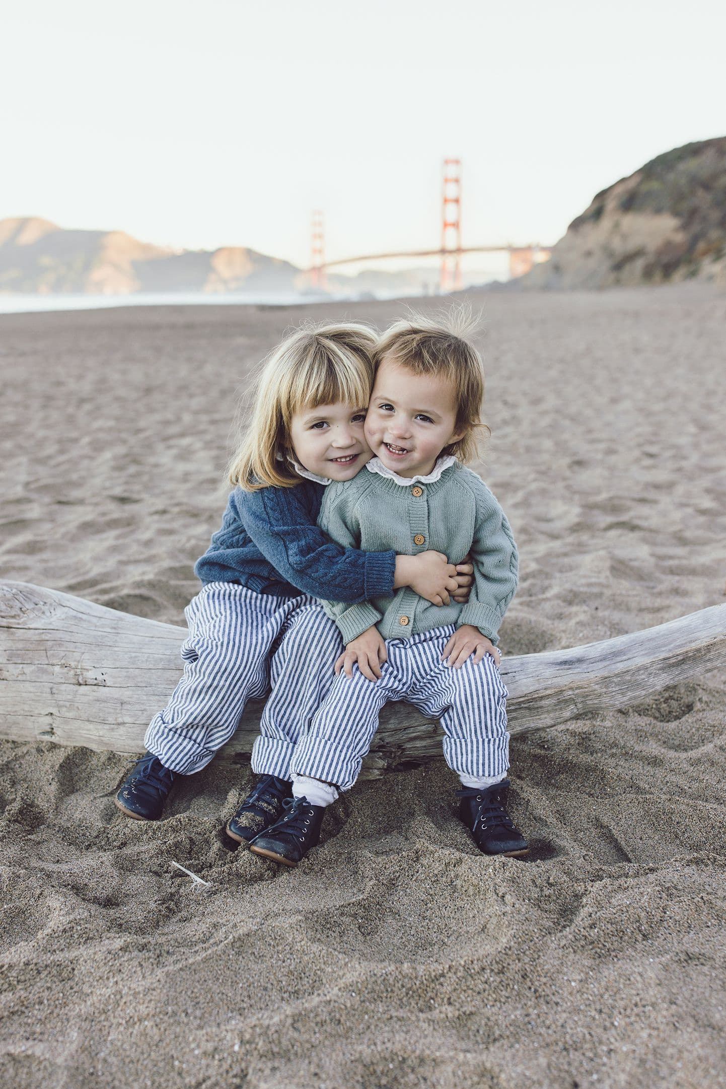 Two small children hugging on a driftwood log at Baker Beach, the Golden Gate Bridge in the haze behind them