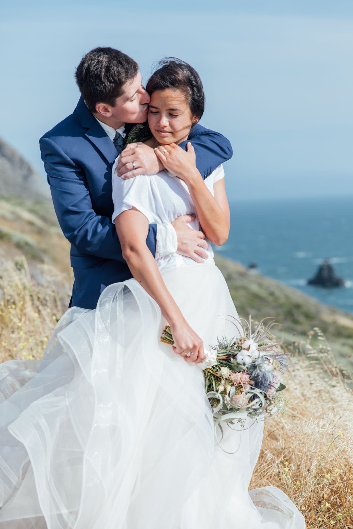 Groom holding his bride from behind on a coastal bluff, her veil and bouquet catching the wind
