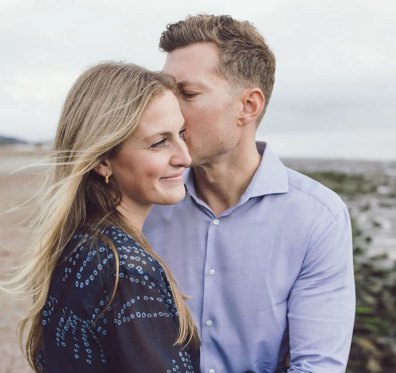 Man kissing his partner's temple on a windy beach