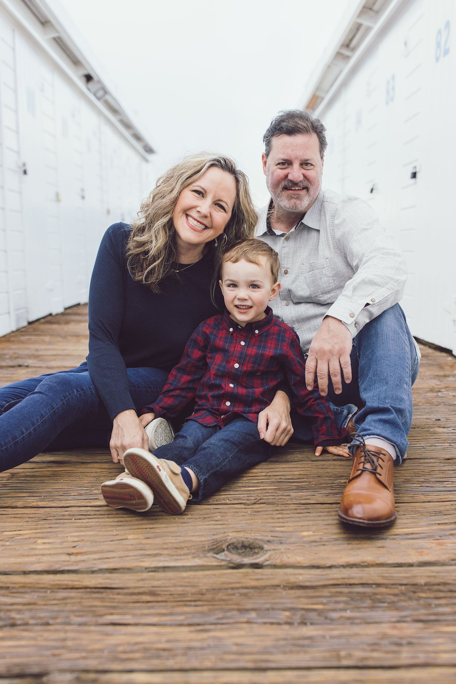 Family of three sitting on a boardwalk in front of white boathouse doors