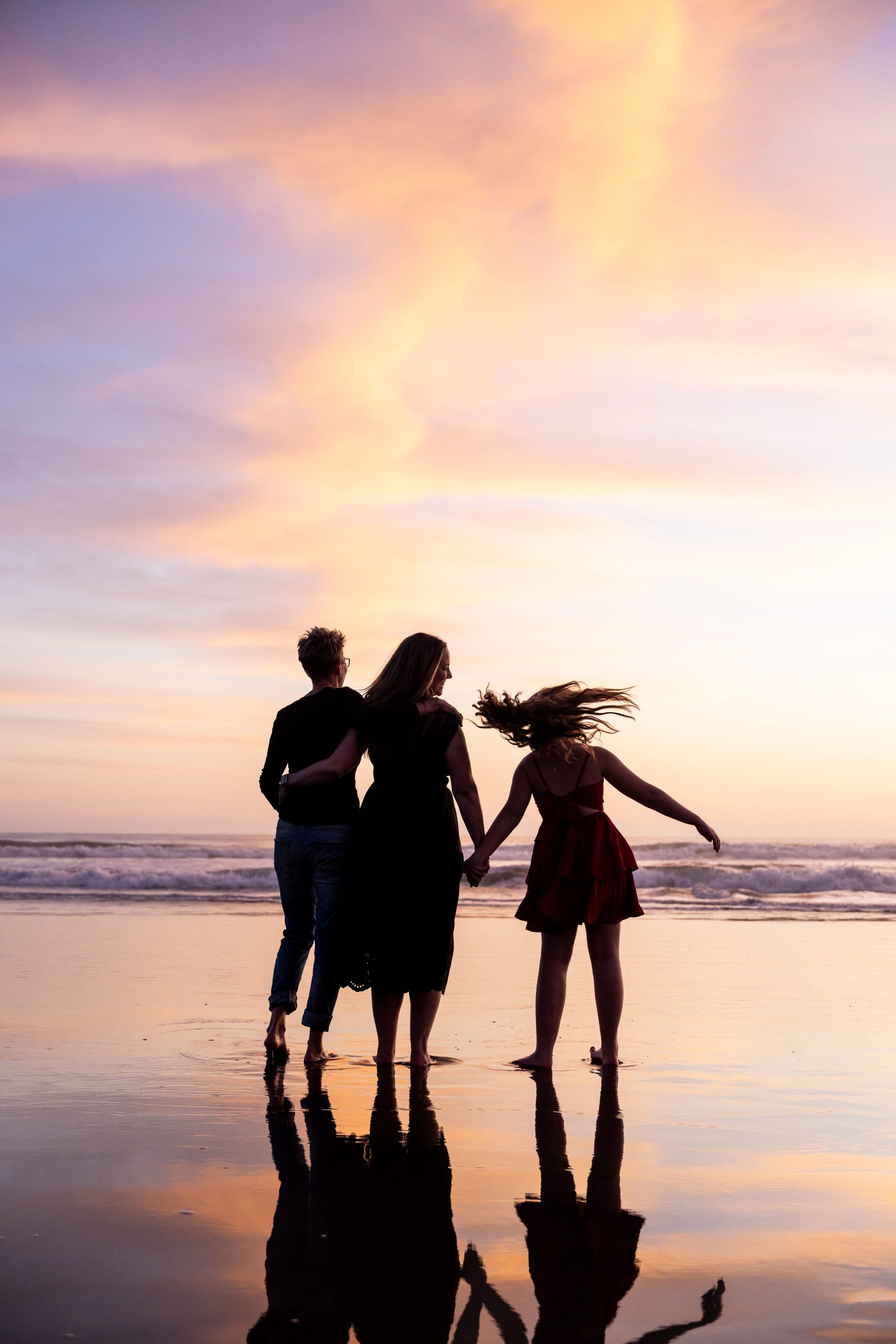 Family of three holding hands at the tideline under a sunset sky