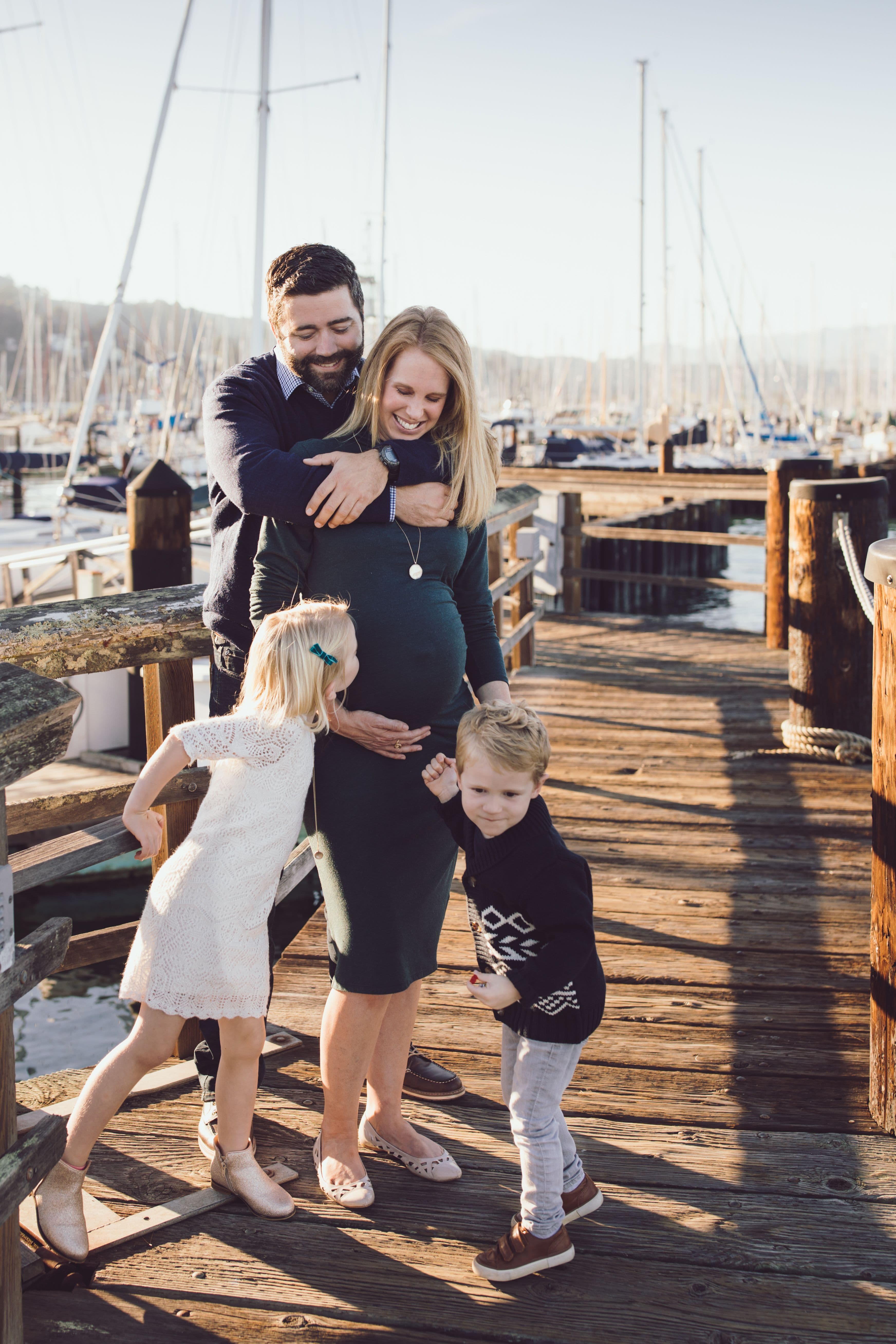 Expectant mother laughing on a marina dock as her family holds on to her