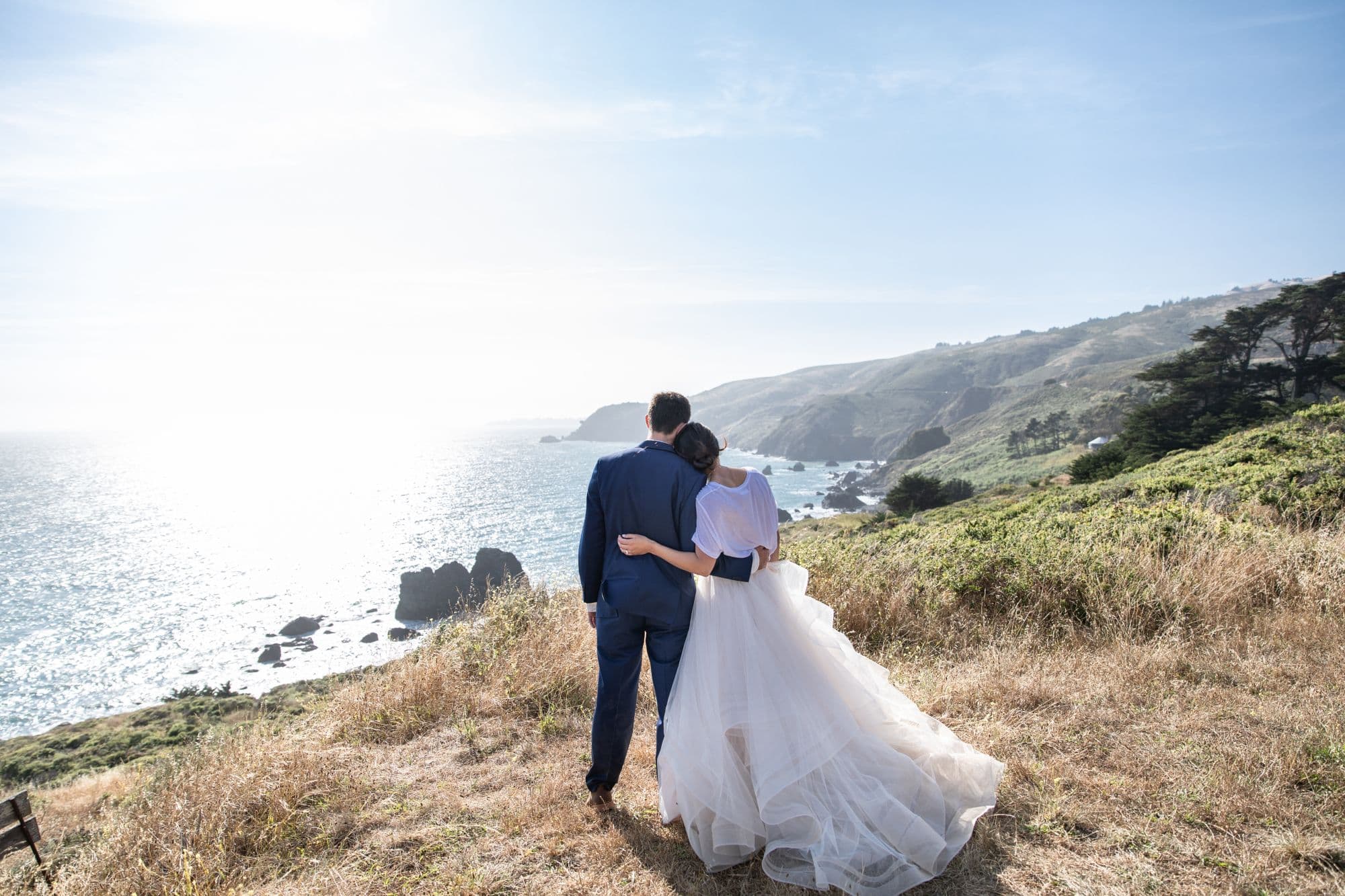 Bride and groom standing arm in arm on a grassy headland looking out over the Pacific
