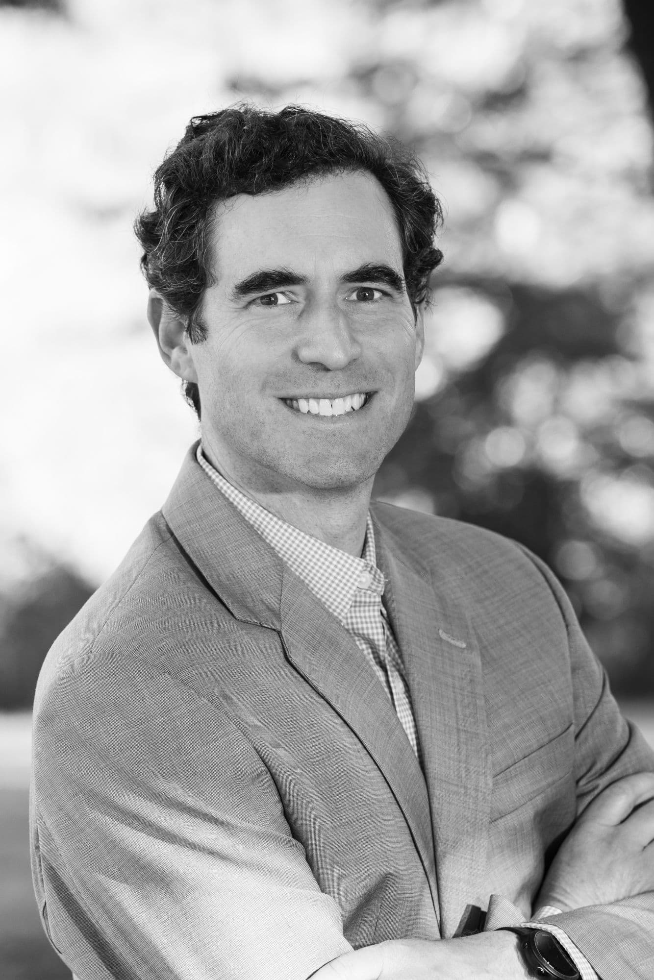 Black-and-white outdoor headshot of a man in a checked shirt and blazer
